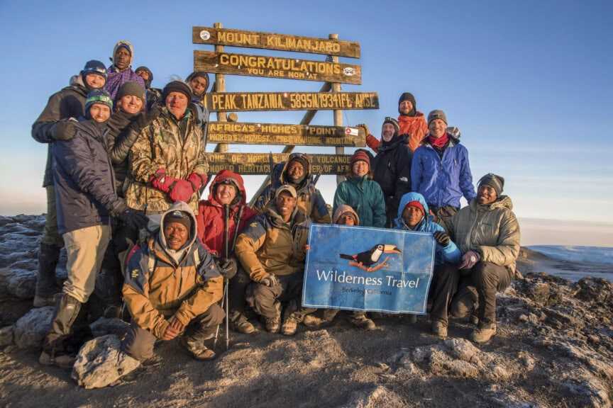 A group of people poses at the Mount Kilimanjaro summit sign, some holding a Wilderness Travel banner, with rocky ground and a distant view behind them—capturing the thrill of this once-in-a-lifetime experience atop Africa’s iconic peak.