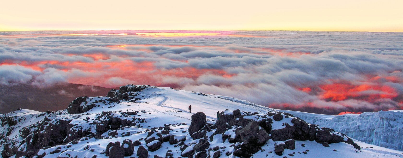 A snowy mountain ridge with a person standing, overlooking a sea of clouds illuminated by a red and orange sunrise, capturing the magic of Kilimanjaro tours.