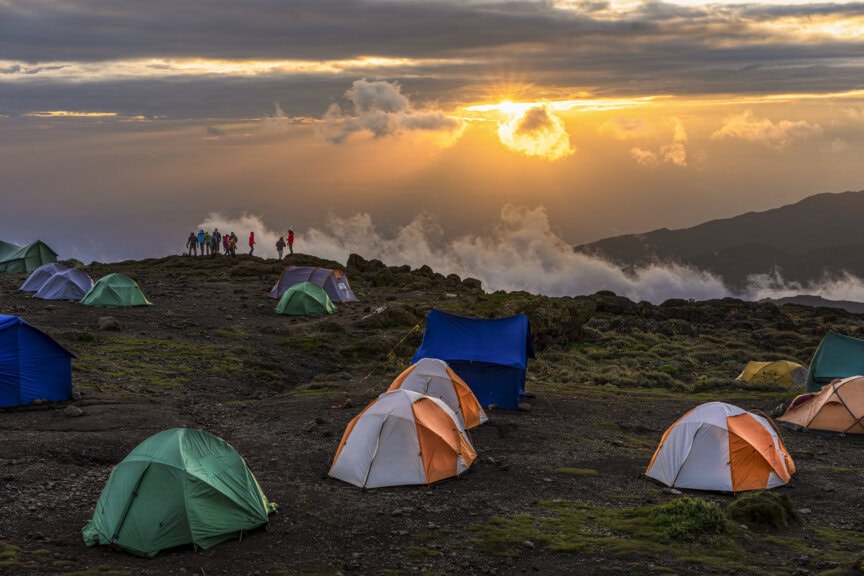 Colorful tents are scattered on a rocky campsite at sunrise, with a group of people standing near the crater’s edge overlooking clouds and distant mountains—a once-in-a-lifetime experience reminiscent of an adventure on Kilimanjaro.