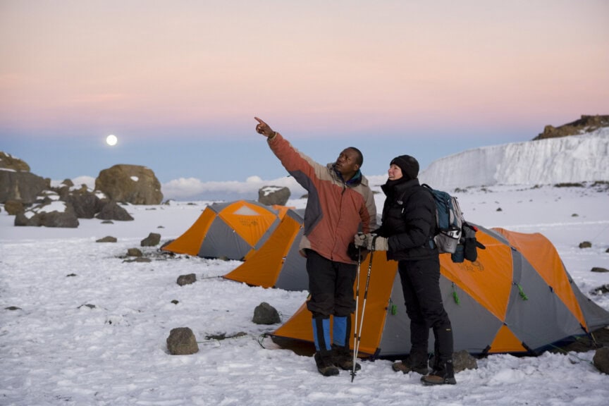Two people in winter gear stand by orange tents on snow, with one pointing toward the sky at dusk. Icy cliffs and rocks are visible in the background, capturing the breathtaking beauty of Kilimanjaro—a once-in-a-lifetime experience for any adventurer.