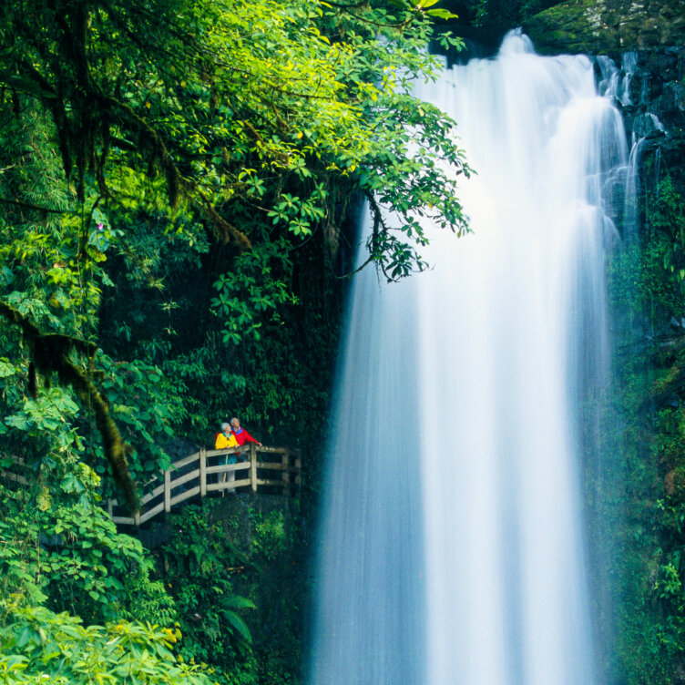 Two people in rain jackets stand on a wooden platform next to a large, flowing waterfall surrounded by dense green foliage—a scene straight out of a Costa Rica travel guide, offering inspiration and tips for exploring the country’s lush natural wonders.
