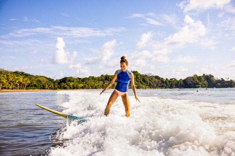 A person in a blue top surfs a small wave near the shore, one of the popular things to do in Costa Rica, with green trees and a sandy beach in the background under a partly cloudy sky—a perfect scene for travel enthusiasts.