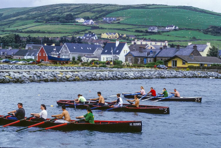 Three rowing teams compete near a rocky shore, with colorful houses and green hills—a perfect scene for Ireland travel, no matter the Ireland weather.