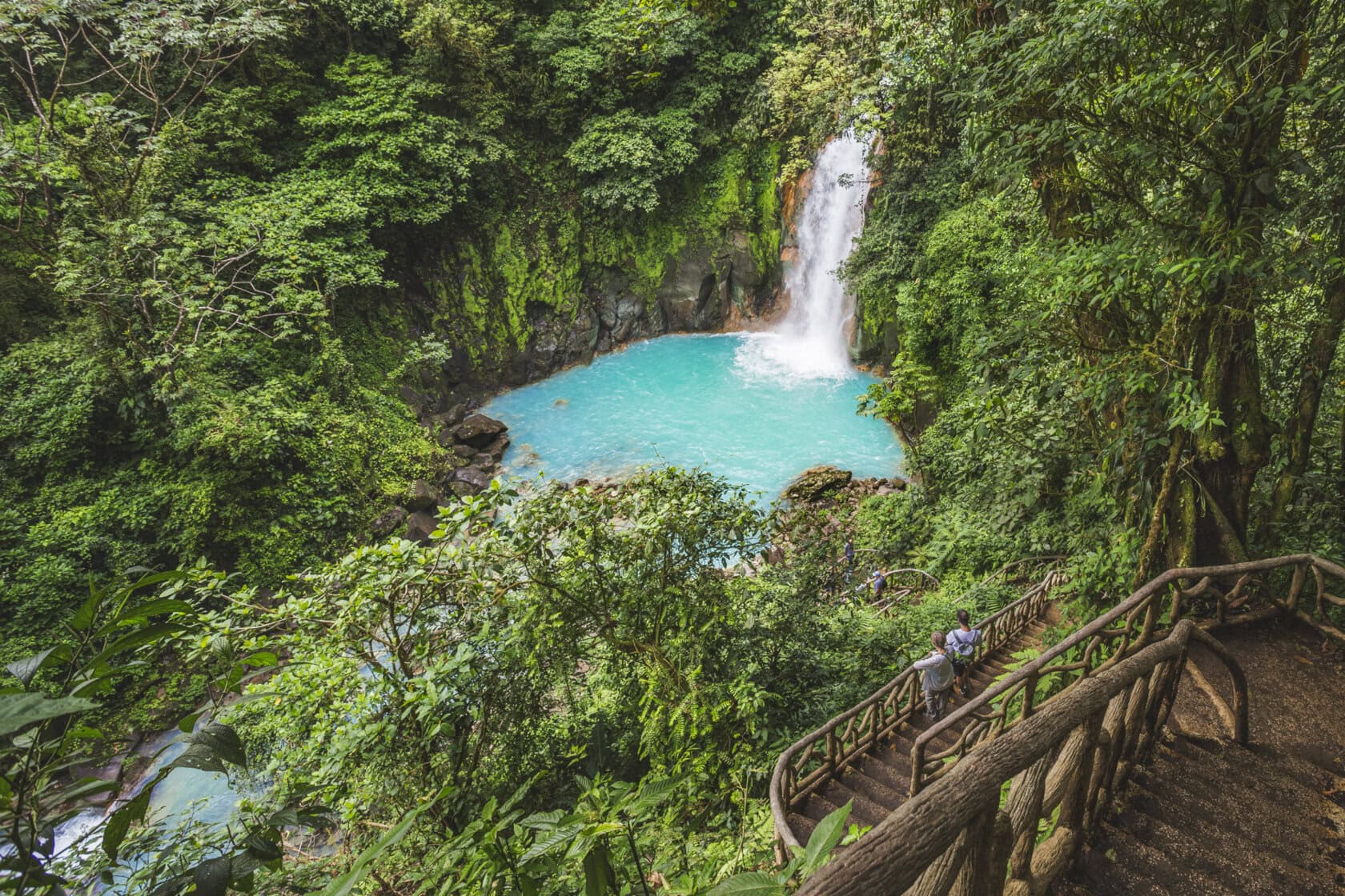 A waterfall flows into a bright turquoise pool surrounded by dense green forest, with a wooden staircase leading down toward the water—a perfect scene for your next Costa Rica trip.