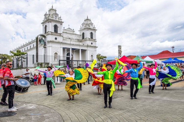 A group of dancers in colorful costumes perform in front of a white church, with musicians and an audience nearby at an outdoor event—a vibrant scene that captures the cultural spirit of Costa Rica and offers visitors a glimpse into local traditions during the best time to visit.