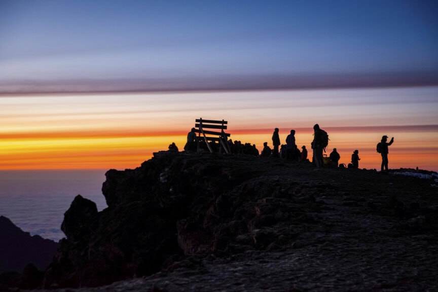 Silhouetted people stand on a rocky peak at sunset, with orange, yellow, and blue layers of sky in the background—capturing the awe of a once-in-a-lifetime experience atop Kilimanjaro’s crater rim.