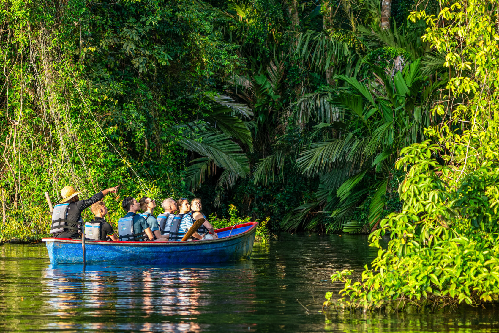 A group of people wearing life jackets sit in a blue boat on a river surrounded by dense green jungle, while a guide points toward the trees in one of the best parks and reserves in Costa Rica.