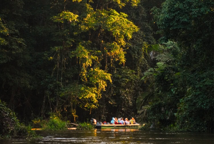 A group of people are seated in a boat on a river surrounded by dense, green jungle, with sunlight illuminating the scene—a perfect way to experience Costa Rica attractions and discover the natural beauty that makes this one of the top things to do in Costa Rica.