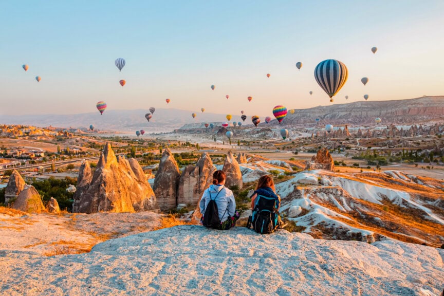 Two people with backpacks sit on a rocky hill, overlooking a valley filled with hot air balloons at sunrise in Cappadocia, Turkey—an experience that truly captures the magic of the best time to visit Turkey.