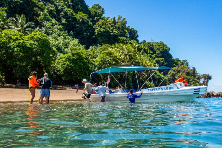 A small group of people boards a white boat named “TAPIR PG8580” from a tropical beach with lush green trees under a clear blue sky—one of the unforgettable things to do while exploring Costa Rica’s vibrant coastline.