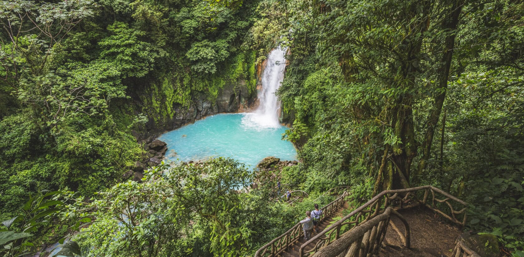 A turquoise waterfall pool surrounded by dense green forest, with a wooden staircase and a person standing on the steps in the foreground—an unforgettable scene that captures the magic of Costa Rica travel.