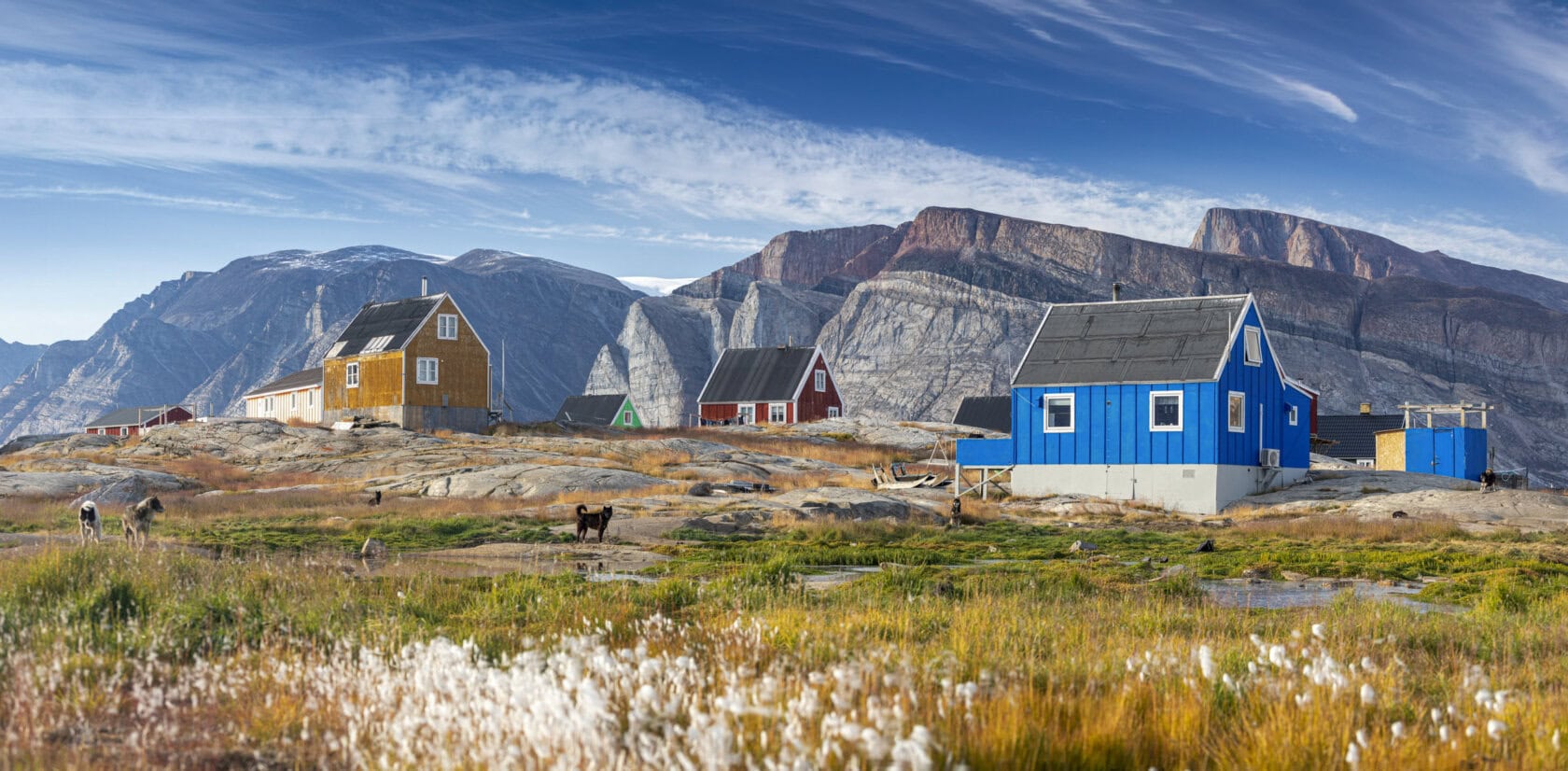 Colorful houses stand on rocky terrain with mountains in the background, grassy vegetation in the foreground, and a blue sky—the best time to visit Greenland is summer.
