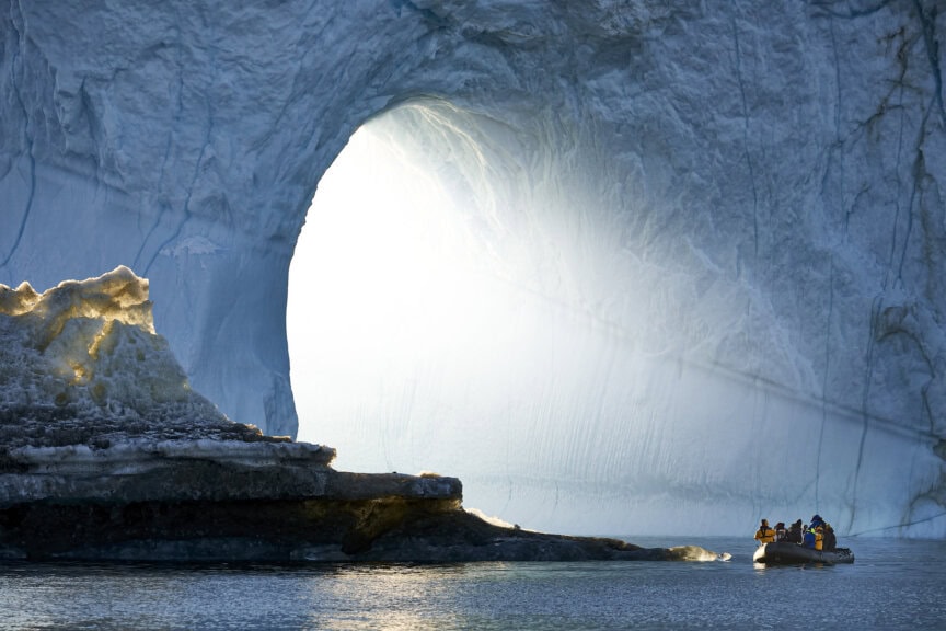 A small boat with people approaches a large ice arch in a glacier, illuminated by sunlight—capturing the magic of the best time to visit Greenland.