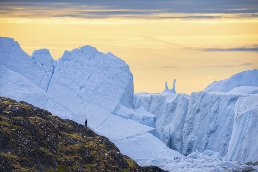 A person stands on a grassy hill overlooking large icebergs under a yellow and orange sky, capturing the best time to visit Greenland.