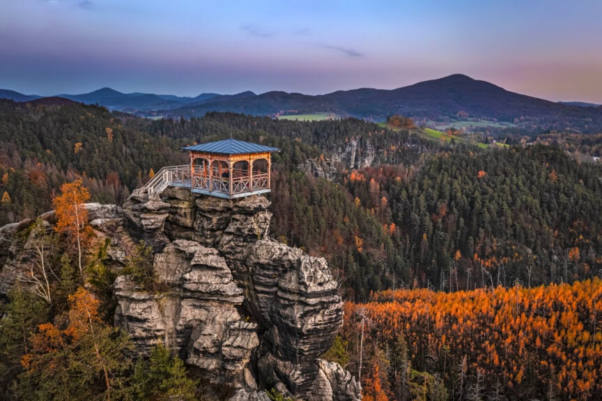 A wooden gazebo with a blue roof sits atop a rocky outcrop, offering stunning views of autumn foliage and distant mountains—an ideal travel spot in the Czech Republic at sunset.