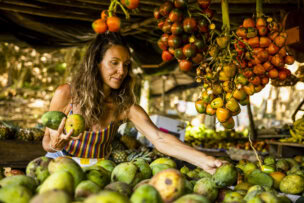 A woman selects mangoes at a market stall in San José, framed by hanging bunches of palm fruit.