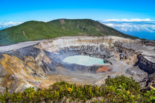 Turquoise volcanic lake inside a wide crater near San José, Costa Rica, surrounded by jagged, rocky terrain and green mountain slopes.