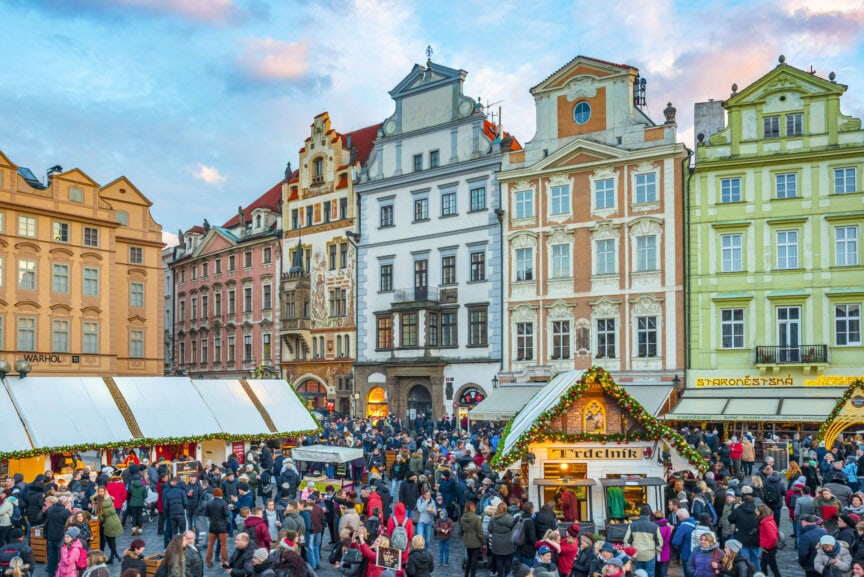 Crowds gather at a festive outdoor market in the Czech Republic, surrounded by colorful historic buildings—an ideal travel spot and the best time to visit under a partly cloudy sky.