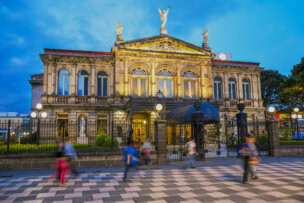 The National Theatre of Costa Rica at twilight, with blurred pedestrians walking on a patterned sidewalk in the foreground of San José.