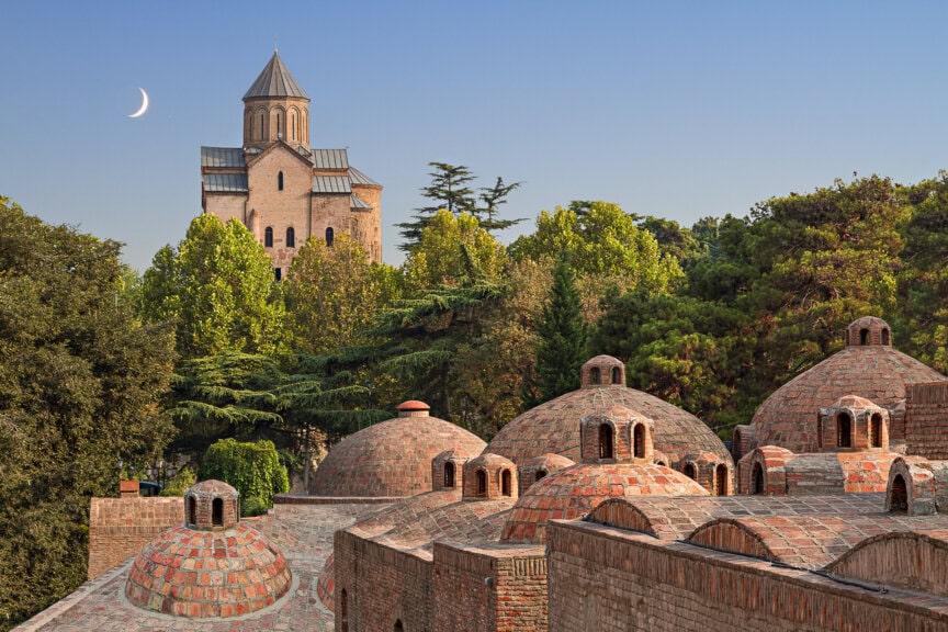 A church with a conical dome sits behind brick bathhouse domes and green trees under a crescent moon, a sight to see during the best time to visit Georgia.