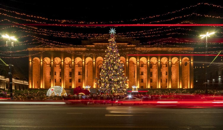 A large, decorated Christmas tree stands before a grand, lit building at night, with light trails from passing traffic reflecting the best time to visit Georgia.
