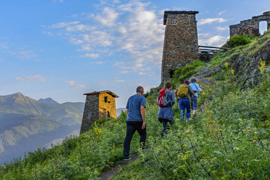Four hikers walk up a grassy mountain path toward two ancient stone towers during the best time to visit Georgia.