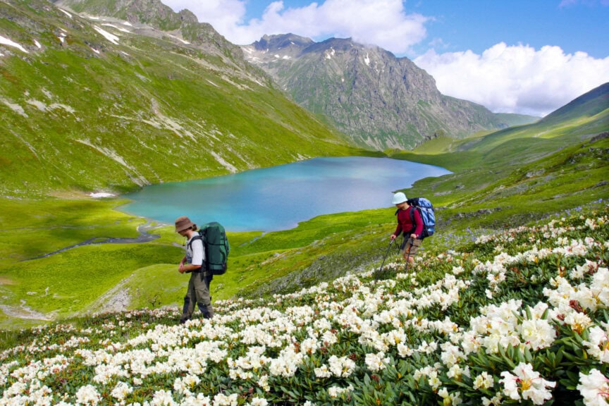 Two hikers with backpacks walk through a field of white flowers in front of a blue mountain lake, an ideal sight during the best time to visit Georgia.